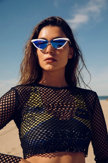 Brunette woman poses on beach wearing sunglasses and a black fishnet crop top against a blue sky, creating a summer fashion portrait.