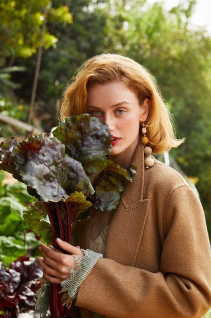 Portrait of a red-haired woman in a tan coat holding beet greens in a garden setting, featuring earthy tones and natural light.