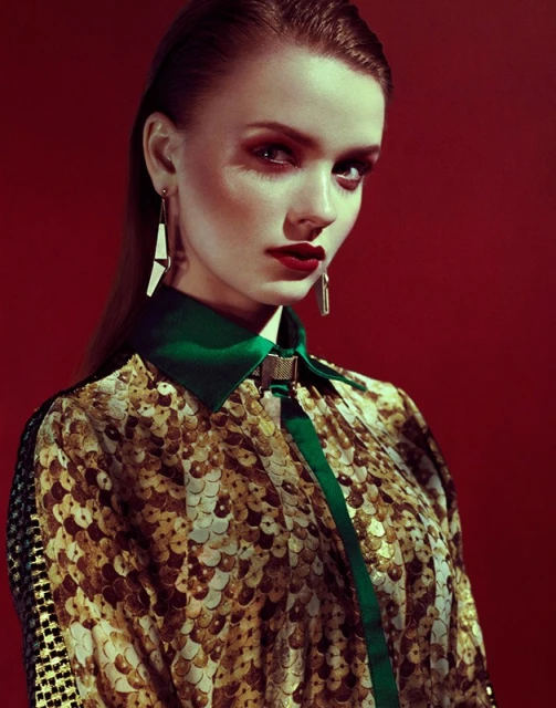 Portrait of a stylish woman with red lips and statement earrings. She wears a patterned top with green trim against a red background.