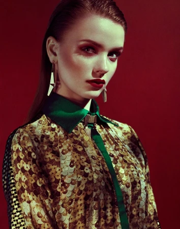 Portrait of a stylish woman with red lips and statement earrings. She wears a patterned top with green trim against a red background.