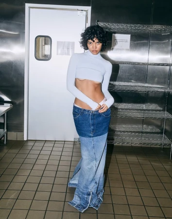 A woman with curly hair poses in a cropped top and a long denim skirt, standing in a room with metal walls and tile floors.