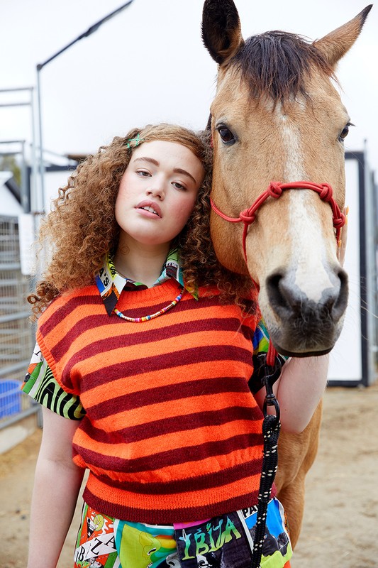 Teen poses with a horse outdoors, wearing an orange striped sweater vest and patterned pants against a neutral-toned outdoor backdrop.