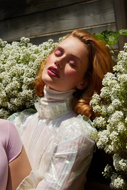 Fair-skinned woman with red hair and pink makeup, eyes closed, surrounded by white flowers with a wood background bathed in sunlight.