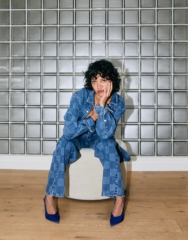 Young woman poses indoors wearing matching checkerboard denim jacket, wide-leg jeans, and heels against a glass block wall backdrop.