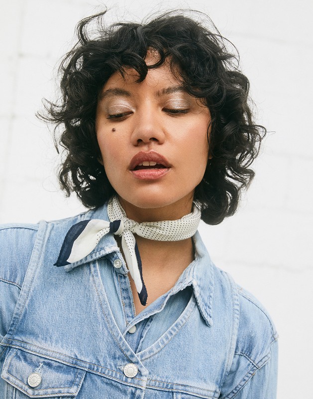 Portrait of an adult woman with curly dark hair wearing a denim jacket and a patterned neck scarf, posing in front of a white brick wall.