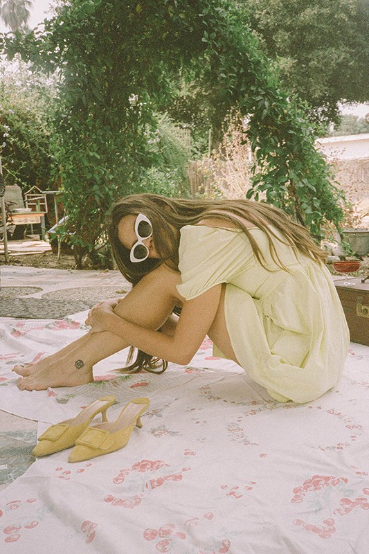 Woman in a pale yellow dress and white cat-eye sunglasses sits on a patterned blanket in a sun-drenched garden setting.