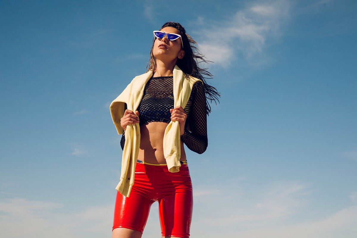 Woman in a black mesh top and red shorts poses outdoors with a yellow sweater around her shoulders against a blue sky background.