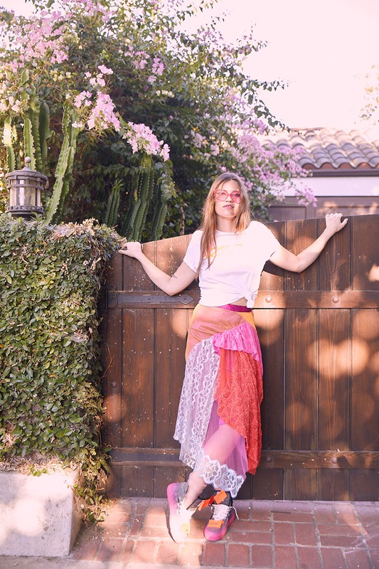 A woman with long hair wearing a white t-shirt and a colorful skirt posing in front of a wooden gate surrounded by greenery and flowers.