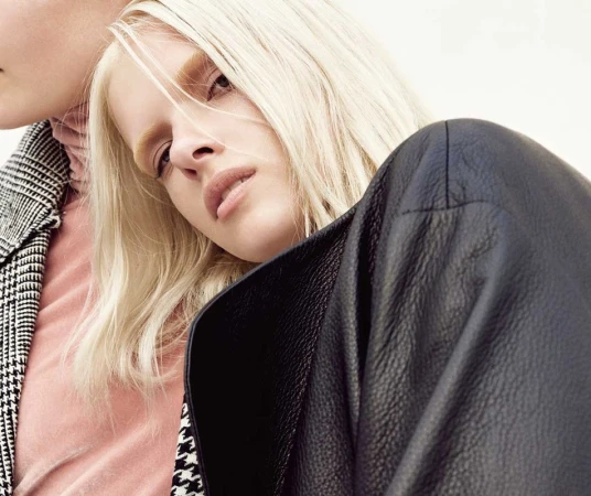 Close-up portrait of a blonde model wearing a black leather jacket with a second model visible in a houndstooth coat against a plain background