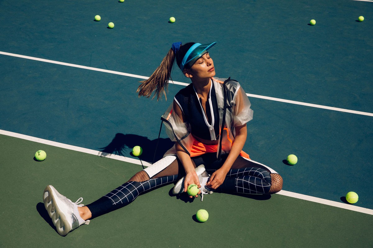 Woman in sporty outfit sits on a tennis court with tennis balls scattered around, wearing a visor and holding a tennis ball.