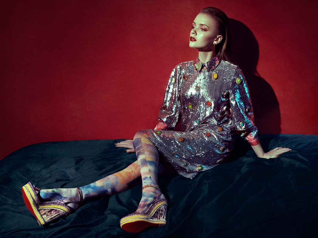 A young woman in a shiny dress and patterned tights poses on a green velvet couch in front of a deep red wall in a fashion shot.