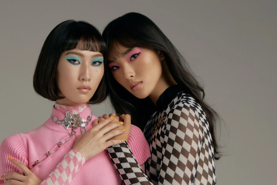 Two Asian fashion models pose in a studio setting wearing pink and black and white patterned clothing with bright eye makeup.