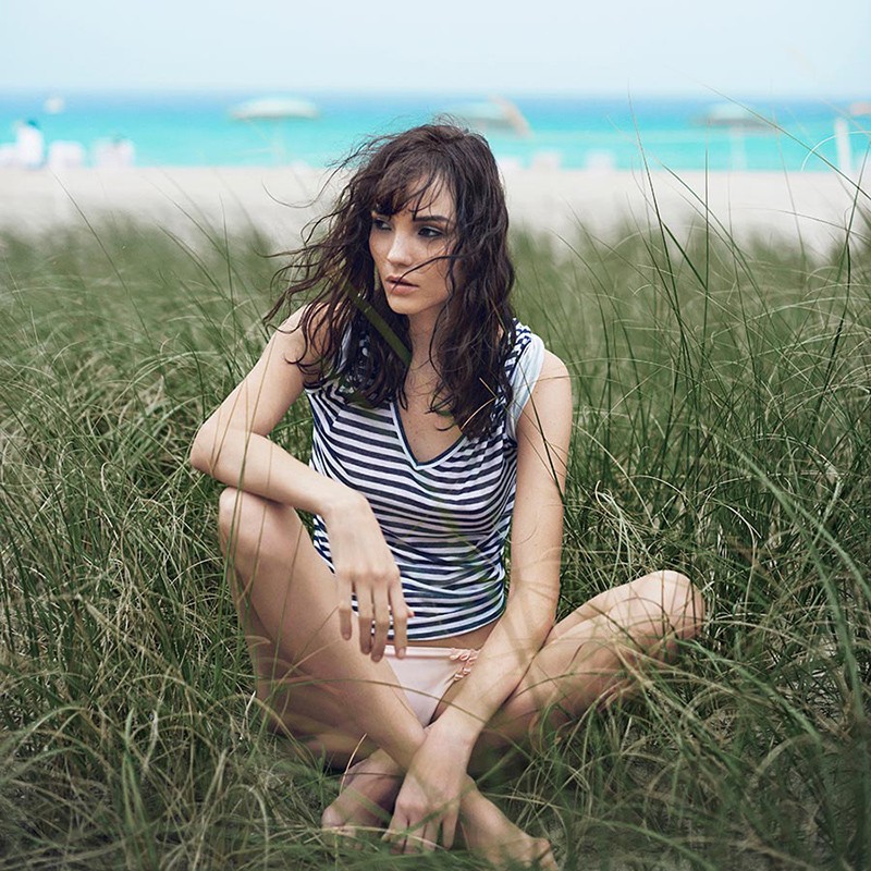 Woman sits in beach grass on a sunny day, wearing a striped shirt, with blurred beach and ocean in the background in outdoor shot.