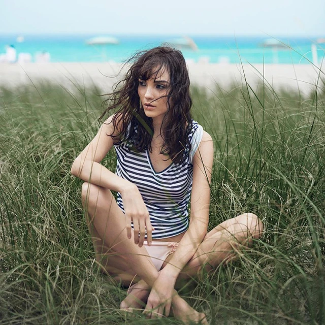 Woman sits in beach grass on a sunny day, wearing a striped shirt, with blurred beach and ocean in the background in outdoor shot.