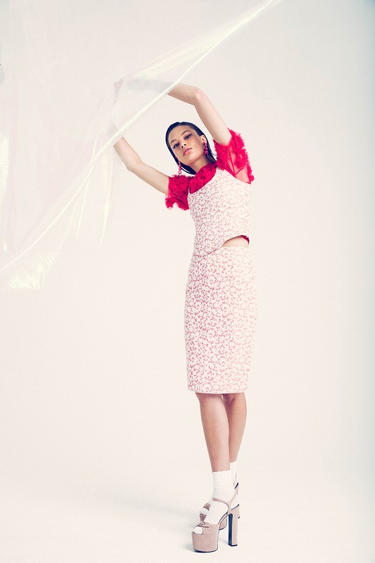 A woman poses in a studio wearing a printed top and skirt with platform heels and a translucent plastic sheet draped above her.