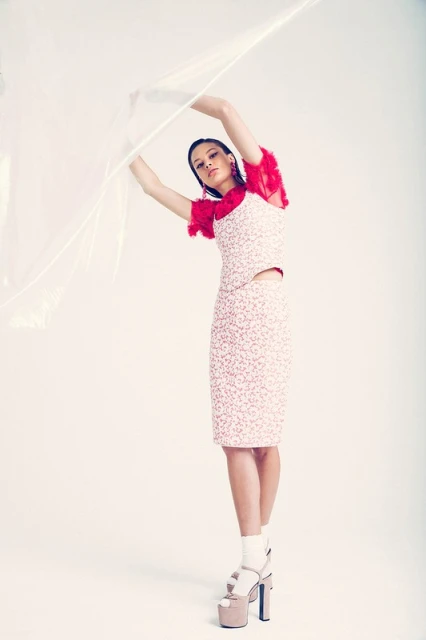 A woman poses in a studio wearing a printed top and skirt with platform heels and a translucent plastic sheet draped above her.
