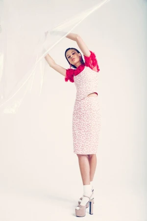A woman poses in a studio wearing a printed top and skirt with platform heels and a translucent plastic sheet draped above her.