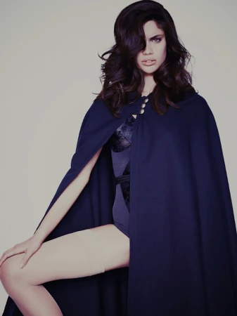 A woman wearing a blue cape and black bodysuit is posing against a neutral background in a studio shot with soft lighting and dark hair.
