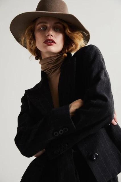 Elegant woman in a pinstripe blazer and wide-brimmed hat, gazing at the camera with red lipstick and neutral scarf, indoor studio setting.
