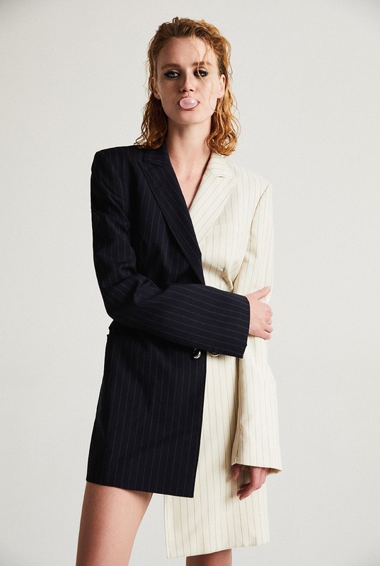 Fashion-forward young adult woman with curly red hair wearing black-and-white pinstripe blazer-dress, blowing bubblegum on a plain light background.