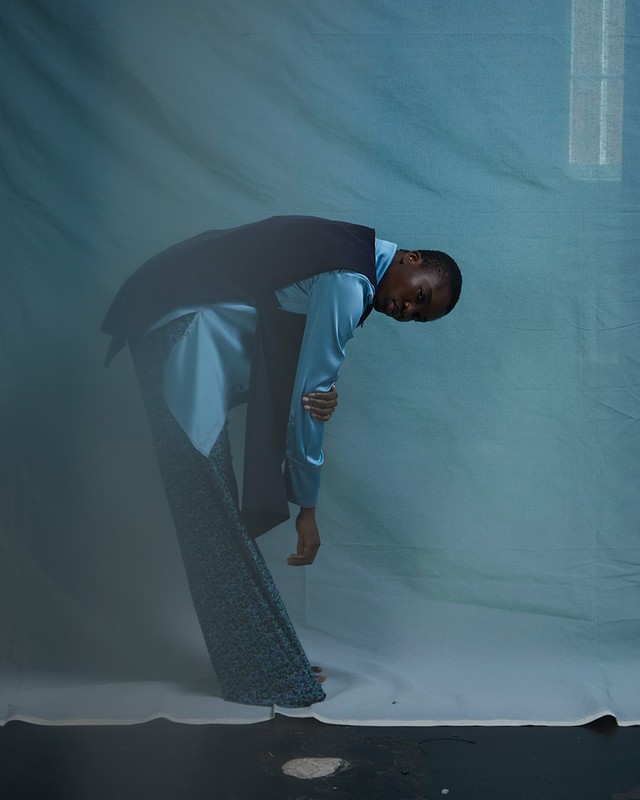 Fashionable adult leans and looks at viewer in blue satin shirt, dark vest, and patterned pants against a mottled blue studio backdrop.