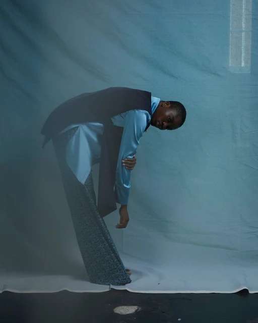 Fashionable adult leans and looks at viewer in blue satin shirt, dark vest, and patterned pants against a mottled blue studio backdrop.