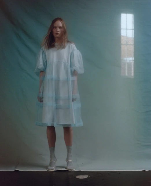 A woman in a light blue dress and grey socks stands in a room with a faded blue background and light coming through a window.