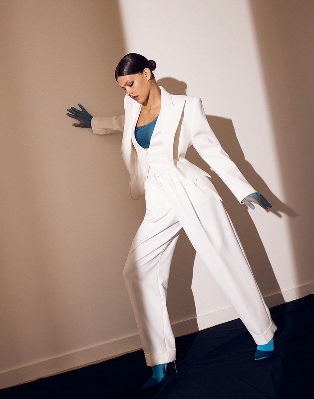 Elegant woman in a white pantsuit strikes a pose against a neutral background, with dramatic shadows enhancing the fashion aesthetic.