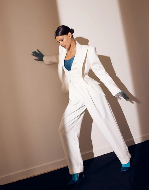 Elegant woman in a white pantsuit strikes a pose against a neutral background, with dramatic shadows enhancing the fashion aesthetic.