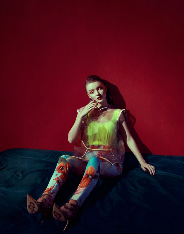 A woman in floral leggings and a sheer top sits on a teal surface against a solid red background in a fashion style image.