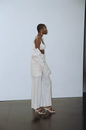 Black woman with short hair wearing a white top and trousers against a pale background