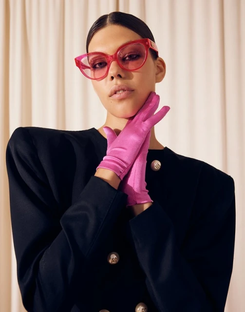 A woman poses in a black double-breasted blazer with pink gloves and pink cat-eye glasses against a neutral-toned curtain backdrop.