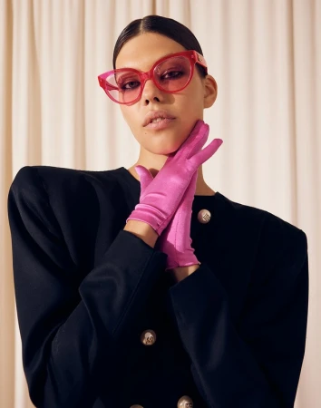 A woman poses in a black double-breasted blazer with pink gloves and pink cat-eye glasses against a neutral-toned curtain backdrop.