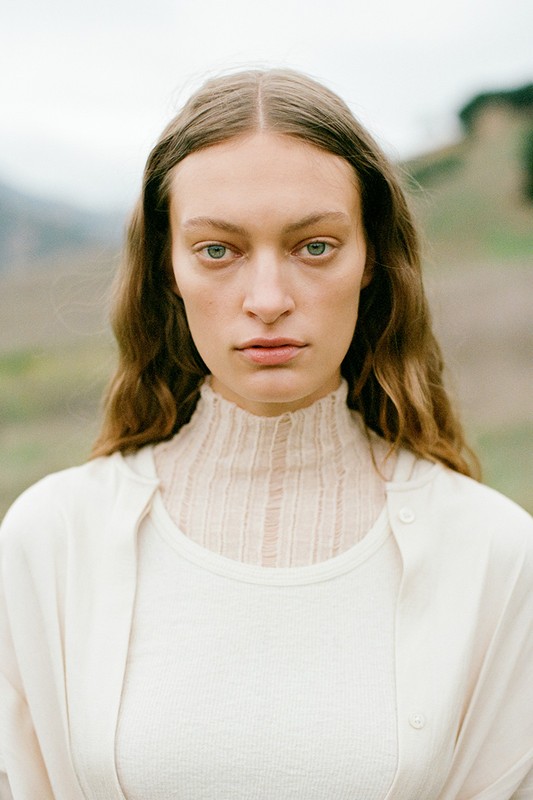 Close-up portrait of an adult female with long brown hair wearing a white shirt and sweater standing outdoors in a rural setting.