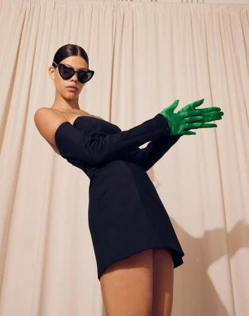 Model in black strapless minidress, heart sunglasses, and green gloves against draped fabric