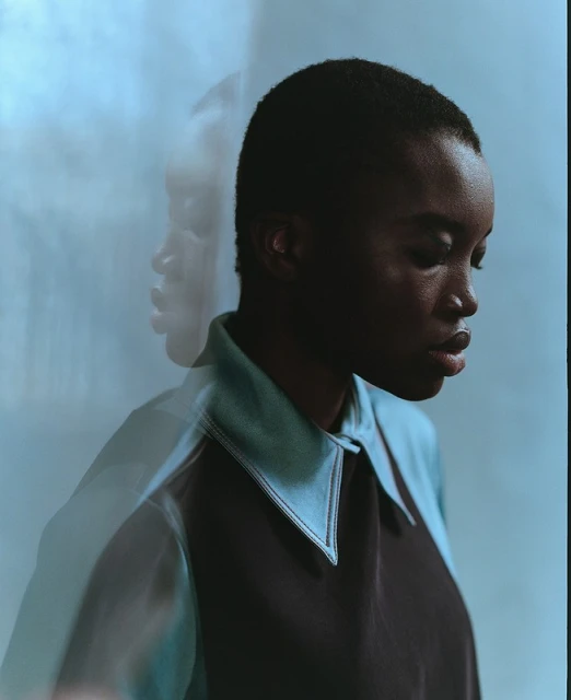 Close-up studio portrait of a young Black woman with short hair, wearing a dark blue and light blue collared shirt against a light blue background.
