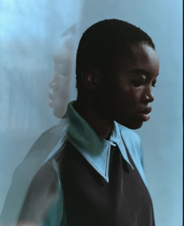 Close-up studio portrait of a young Black woman with short hair, wearing a dark blue and light blue collared shirt against a light blue background.