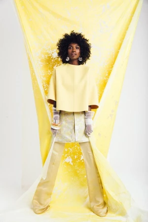 A woman with an afro hairstyle poses in a yellow cape top and shiny gold pants against a bright backdrop for a fashion editorial.