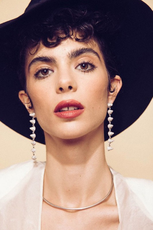 Close-up portrait of a woman wearing a black wide-brimmed hat, long drop earrings, and a metallic choker necklace.