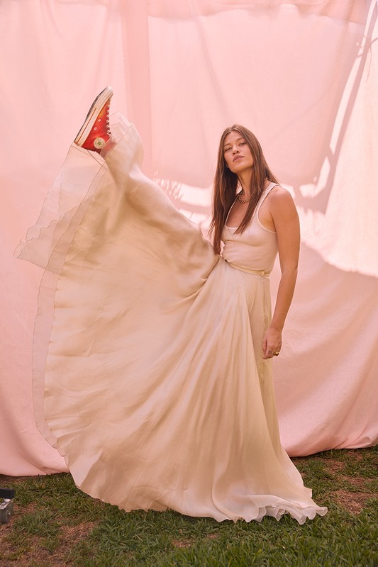 Woman in flowing beige dress and red sneaker poses in front of a soft pink backdrop, one leg raised, for fashion-editorial image.
