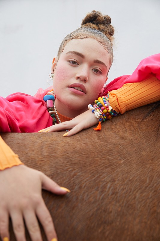Portrait of a young adult woman wearing a pink jacket, orange top, and beaded jewelry, leaning against a brown horse in outdoor setting.