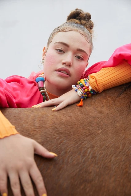 Portrait of a young adult woman wearing a pink jacket, orange top, and beaded jewelry, leaning against a brown horse in outdoor setting.