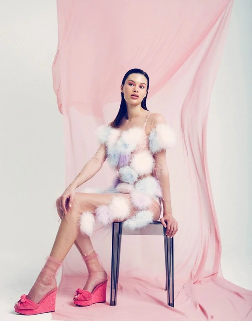Brunette model poses seated on stool in feathered mini-dress and pink platforms against a pink-and-white backdrop in a studio shot.
