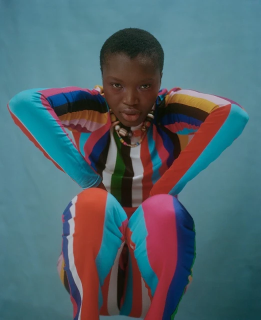 Fashion portrait of an African woman in a colorful striped outfit against a pale blue background, posing in a studio setting with a necklace.