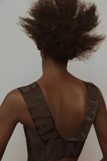 Back view of a woman with afro hair wearing a brown top with a large letter pattern against a neutral background in an indoor setting.