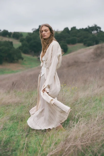 Woman in flowing beige dress stands in grassy hills with trees under cloudy sky, looking at the camera in a fashion editorial.