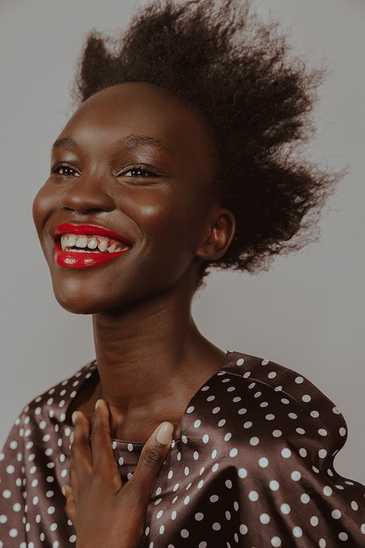 Close-up of a smiling African woman with red lipstick wearing a polka dot top against a neutral background in a beauty shot.