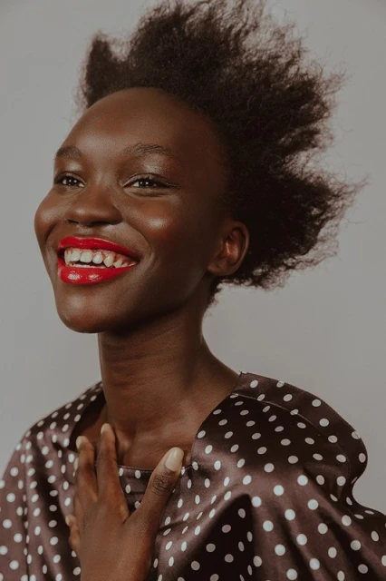 Close-up of a smiling African woman with red lipstick wearing a polka dot top against a neutral background in a beauty shot.