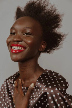 Close-up of a smiling African woman with red lipstick wearing a polka dot top against a neutral background in a beauty shot.