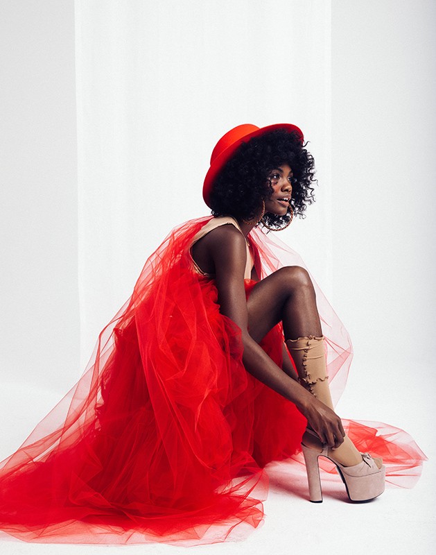 Woman in red tulle dress and matching hat adjusting high-heeled shoe in a photo studio. Beauty and fashion photography.
